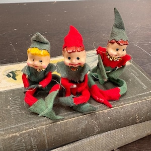 Puede incluir: Tres figuras de elfos vintage con caras pintadas, trajes de fieltro rojo y verde y sombreros puntiagudos. Los elfos están sentados en una pila de libros viejos. Cada elfo tiene un collar dorado y zapatos rojos.