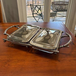 以下が含まれることがあります： 2つのコンパートメントと蓋が付いたステンレス製のサービングディッシュ。竹風のハンドルが付いており、木製のテーブルの上に置かれています。蓋には小さな装飾的なハンドルが付いています。