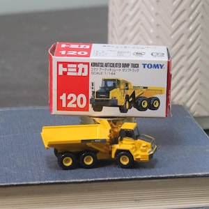 Pode incluir: Um brinquedo de caminh&atilde;o basculante articulado Komatsu amarelo com rodas pretas e cabine preta. O brinquedo &eacute; um modelo em escala, com a caixa lendo "Scale 1/144". A caixa &eacute; vermelha e branca com texto japon&ecirc;s.