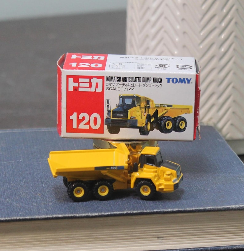 Pode incluir: Um brinquedo de caminh&atilde;o basculante articulado Komatsu amarelo com rodas pretas e cabine preta. O brinquedo &eacute; um modelo em escala, com a caixa lendo "Scale 1/144". A caixa &eacute; vermelha e branca com texto japon&ecirc;s.