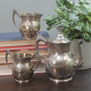 Puede incluir: Un juego de té plateado con detalles florales. El juego incluye una tetera, una crema y un azucarero. La tetera tiene un pico curvo y una tapa decorativa. La crema y el azucarero tienen asas ornamentadas.