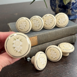 May include: A set of cream ceramic cabinet knobs with a floral pattern. Each knob has a white flower with green leaves and a silver border. The knobs are round and come in various sizes, suitable for furniture.