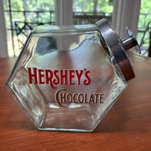 Può includere: Un barattolo di vetro trasparente, esagonale, con coperchio e manico argentati. Il barattolo presenta il logo Hershey's Chocolate in lettere rosse e marroni. Il barattolo è vuoto e poggia su una superficie di legno.