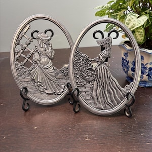 Può includere: Due placche decorative ovali color argento con supporti in metallo nero. Ogni placca presenta un rilievo dettagliato di una donna in un abito lungo, una vicino a un traliccio e l'altra con un cesto di fiori. Le placche sono esposte su una superficie di legno scuro.