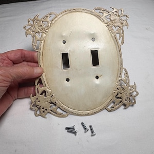May include: Off-white, oval-shaped light switch cover with two switch openings. The cover has decorative floral accents and includes four screws. The cover is held by a hand.