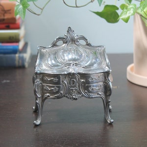 Op de afbeelding: Een kleine, sierlijke zilverkleurige sieradendoos met twee lades en een decoratief bovenstuk. De doos heeft gebogen poten en ingewikkelde details. De doos staat op een donkere houten ondergrond.