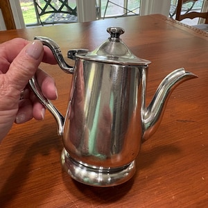 Puede incluir: Una tetera de metal plateado pulido con cuerpo cilíndrico, asa curva, tapa con bisagra y un pequeño pomo, y un pico vertedor cónico. La superficie de la tetera es lisa y reflectante, mostrando un brillo metálico en todo el diseño.