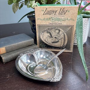 May include: A silver heart-shaped serving dish with a spoon, part of a "Laurel Mist" party set. The dish has a decorative floral border and is next to a box with the same design. The box reads "For the young at heart."