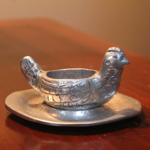 May include: A silver-colored metal chicken-shaped candle holder. The bird has a detailed textured design and sits on a small, round plate. The candle holder is designed to hold a small candle.