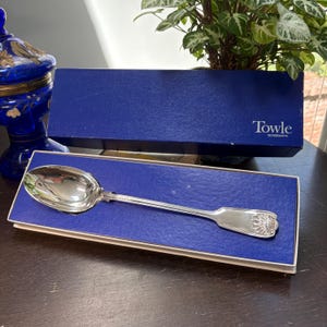 Puede incluir: Una cuchara de plata con un mango adornado descansa en una caja azul. La caja tiene el nombre "Towle Silversmiths" impreso. La cuchara es muy reflectante y parece estar hecha de metal pulido.
