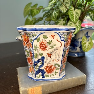 Op de afbeelding: Een vierkante keramische plantenbak met een blauwe rand en handvatten. De plantenbak heeft een handgeschilderd ontwerp van oranje bloemen, groene bladeren en een lichtbruin dier. De plantenbak staat op een boek.