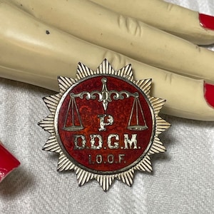 Könnte beinhalten: Eine Vintage-Anstecknadel mit einem Sternmuster. Die Anstecknadel hat eine rote Mitte mit den Buchstaben "P O.D.G.M. I.O.O.F." und Waagen der Gerechtigkeit. Die Anstecknadel hat einen silberfarbenen Rand.