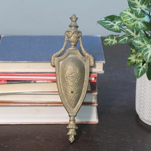 May include: Ornate brass wall sconce with a shield-shaped design. The sconce features detailed floral and scrollwork, with a decorative finial at the top and a tassel-like element at the bottom. The brass has a warm, aged patina.