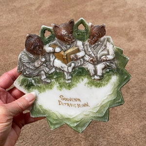 May include: A ceramic souvenir plate featuring three anthropomorphic bears in suits, reading a book. The plate has a scalloped edge with green accents and the text "Souvenir Detroit, Minn." in gold lettering.