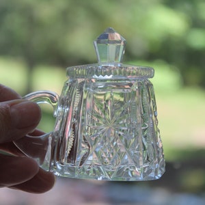 May include: A clear, cut-glass sugar bowl with a lid and handle. The bowl features a starburst pattern on the base and sides, with a diamond-cut design. The lid has a faceted, pointed finial. The handle is a curved loop.