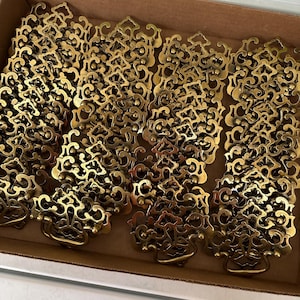 Peut inclure: Une collection de poignées de tiroir en laiton présentant un design orné et symétrique avec des volutes, un point de fixation central et une poignée tombante articulée. Ces pièces métalliques sont rangées dans un carton.
