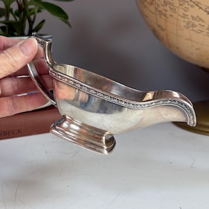 May include: A silver gravy boat with a handle and a decorative rim. The boat has a curved spout and sits on a rectangular base. The gravy boat is likely vintage or antique.