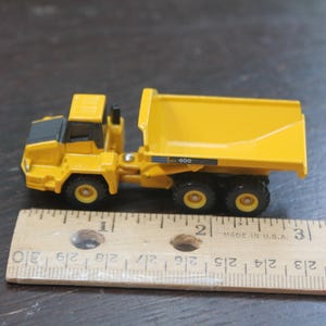 Pode incluir: Um cami&atilde;o basculante de brinquedo amarelo com detalhes em preto e seis rodas. O cami&atilde;o tem uma caixa basculante e est&aacute; ao lado de uma r&eacute;gua de madeira. O texto "400" &eacute; vis&iacute;vel na lateral do cami&atilde;o.