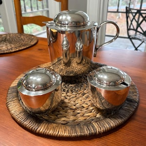 May include: A polished silver tea set, including a teapot with a curved handle and two small lidded bowls, sits on a woven tray. The set reflects the surrounding environment, creating a mirrored effect. The tray is placed on a wooden table.