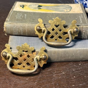 Puede incluir: Dos tiradores de cajón antiguos de latón con diseños calados ornamentados. Cada tirador presenta una placa trasera decorativa y un asa curvada. Los tiradores se colocan sobre un fondo de libros antiguos, lo que sugiere un tema vintage o reciclado.