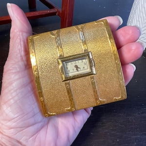 Puede incluir: Un estuche cuadrado de color dorado con una pequeña esfera de reloj. El estuche tiene una superficie texturizada con detalles metálicos decorativos y un reloj rectangular con números negros. El reloj muestra la hora 7:00.