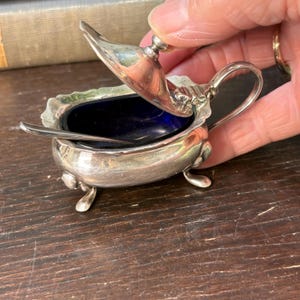 May include: A small, antique silver salt cellar with a hinged lid and a small spoon. The interior is a deep blue color. The salt cellar has three small feet and a handle. The lid is open, revealing the interior.