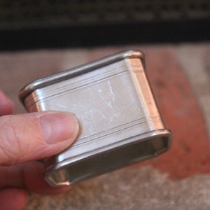 May include: A rectangular silver napkin ring with a slightly curved shape. The ring has a polished finish with subtle horizontal lines and a simple, elegant design. The item is held in a hand.