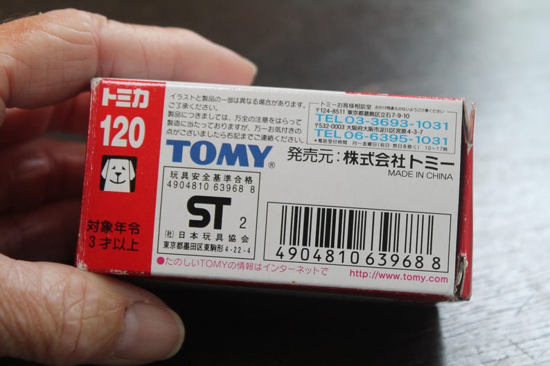 Pode incluir: Uma caixa de brinquedos Tomy vermelha e branca com texto japon&ecirc;s e um logotipo de cachorro. A caixa exibe o n&uacute;mero 120 e o texto "Made in China". A caixa &eacute; segurada por uma m&atilde;o.