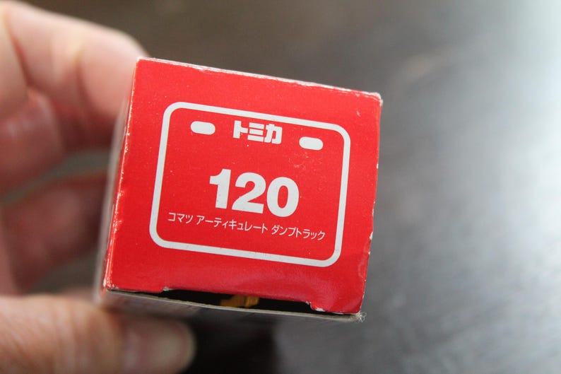 Pode incluir: Uma caixa vermelha com texto e gr&aacute;ficos brancos. O texto inclui o n&uacute;mero "120" e caracteres japoneses. A caixa &eacute; para um ve&iacute;culo de brinquedo, provavelmente um cami&atilde;o basculante.
