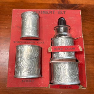 May include: A vintage condiment set in a red box. The set includes four silver-colored containers with a floral design, labeled "Worcestershire Sauce," "Ketchup," and "Mustard." A glass bottle with a black top is also included, labeled "Horse Radish."