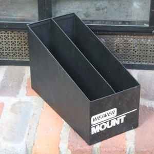 May include: Black metal magazine rack with two compartments. The rack has a rectangular shape and is angled. The text "WEAVER MOUNT" is printed in white on the side of the rack. The rack is sitting on a brick surface.