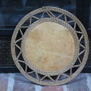 May include: A round, woven basket with a light brown birch bark center. The outer rim features a geometric design, and the basket is made of natural fibers. The basket is likely handmade and suitable for decorative or functional use.
