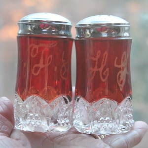 Puede incluir: Un par de saleros y pimenteros de cristal rojo rubí con tapas plateadas. Los saleros y pimenteros tienen una base de cristal tallado transparente y están grabados con una inicial en cursiva. Un complemento clásico para cualquier mesa.