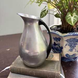Puede incluir: Una jarra plateada vintage con un asa negra. La jarra tiene las palabras "JOHNNIE WALKER" grabadas en la parte delantera. La jarra está colocada encima de una pila de libros antiguos. Una planta en maceta está en el fondo.