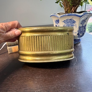 Könnte beinhalten: Ein Vintage-Pflanzgefäß aus Messing mit vertikalen Rippendetails. Der zylindrische Behälter hat einen abgerundeten Boden und einen oberen Rand. Der Pflanztopf hat eine warme Goldfarbe und scheint aus Metall zu sein. Im Hintergrund befindet sich ein blau-weißes Blumenpflanzgefäß.