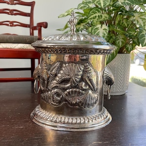May include: A silver-colored, lidded container with seashell and aquatic life relief details. The container has two handles and a decorative finial on the lid. The container is sitting on a dark wooden surface.