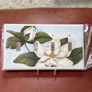 May include: A decorative light switch cover with a watercolour painting of a magnolia flower and bud with green leaves against a light blue background. The cover has three switch openings and is signed by the artist.