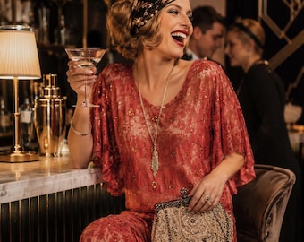 Könnte beinhalten: Ein rotes Spitzenkleid mit kurzen, flatternden Ärmeln und aufwendigen Paillettenverzierungen, kombiniert mit einer goldfarbenen Perlenhandtasche, einer langen Goldhalskette mit Anhänger und einem schwarzen Pailletten-Stirnband mit V-Ausschnitt.