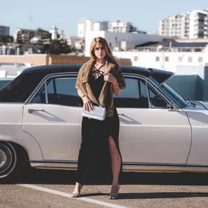 May include: A woman in a black dress and a brown fur shawl stands next to a white vintage car. She is holding a silver clutch purse and is wearing silver heels.