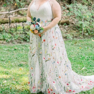 May include: A bride wearing a white wedding dress with colorful floral embroidery. She is holding a bouquet of flowers and smiling at the camera.