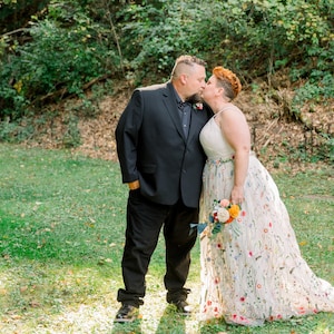 May include: A couple in wedding attire kiss in a grassy outdoor setting. The groom wears a black suit and the bride wears a white dress with colorful floral embroidery.
