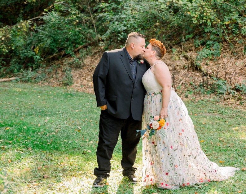 May include: A couple in wedding attire kiss in a grassy outdoor setting. The groom wears a black suit and the bride wears a white dress with colorful floral embroidery.