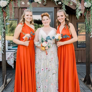 May include: Three people in orange dresses stand in front of a wooden archway decorated with flowers. The person in the middle is wearing a white dress with floral embroidery. They are all holding bouquets of flowers.