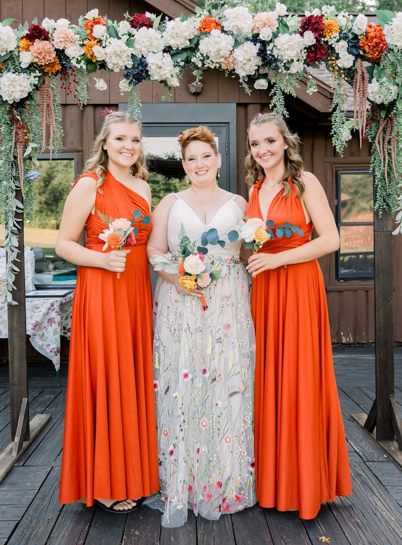 May include: Three people in orange dresses stand in front of a wooden archway decorated with flowers. The person in the middle is wearing a white dress with floral embroidery. They are all holding bouquets of flowers.