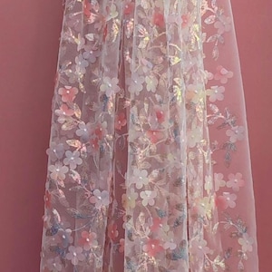 May include: A sheer, white fabric adorned with three-dimensional floral embellishments in pastel pink, yellow, and blue. The delicate flowers and leaves create a textured, romantic aesthetic, ideal for bridal or special occasion wear.