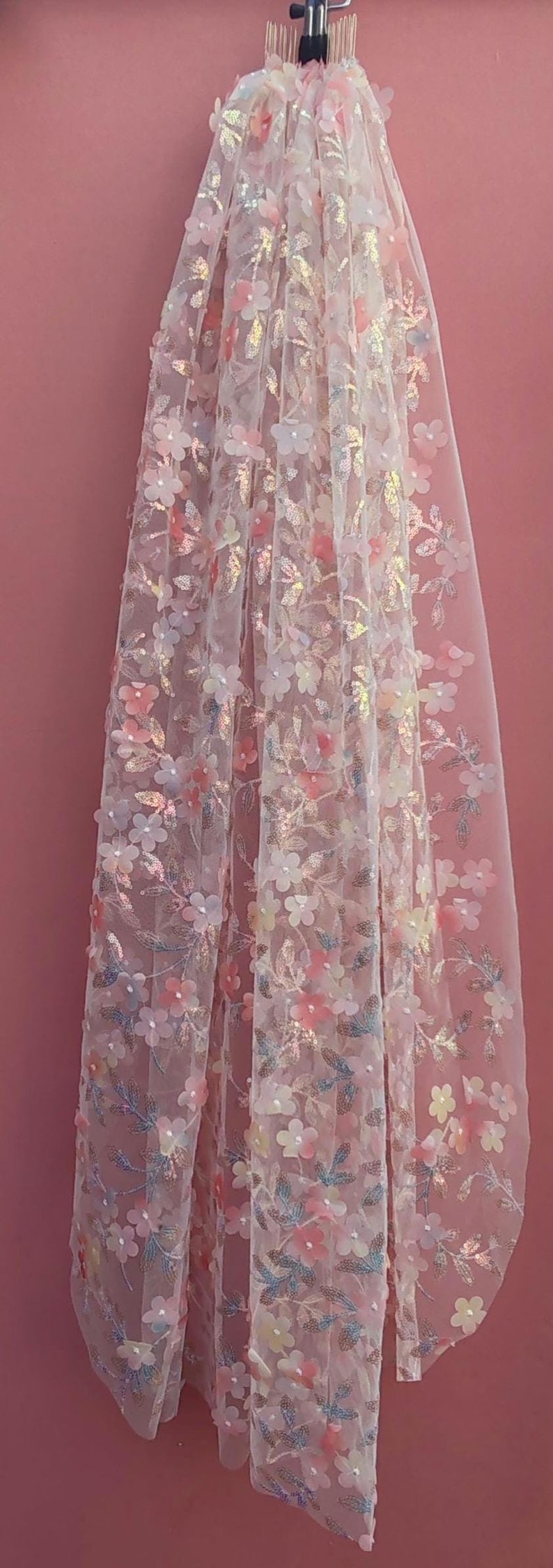 May include: A sheer, white fabric adorned with three-dimensional floral embellishments in pastel pink, yellow, and blue. The delicate flowers and leaves create a textured, romantic aesthetic, ideal for bridal or special occasion wear.