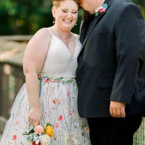May include: A bride and groom are smiling at each other. The bride is wearing a white dress with colorful floral embroidery and a bouquet of flowers. The groom is wearing a black suit and a boutonniere.