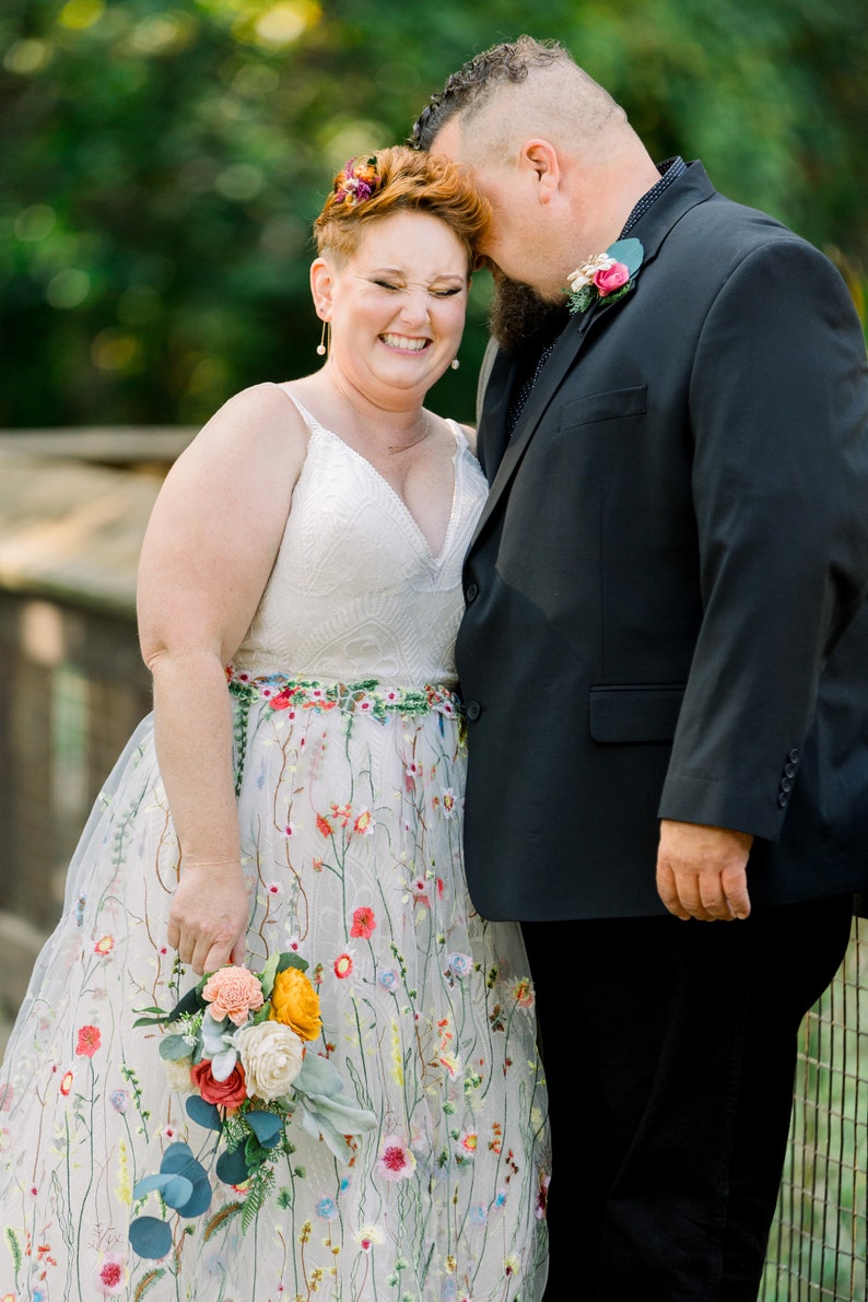 May include: A bride and groom are smiling at each other. The bride is wearing a white dress with colorful floral embroidery and a bouquet of flowers. The groom is wearing a black suit and a boutonniere.
