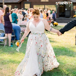 May include: A bride and groom walking down the aisle at an outdoor wedding ceremony. The bride is wearing a white dress with floral embroidery and carrying a bouquet of orange and yellow flowers. The groom is wearing a black suit and tie.