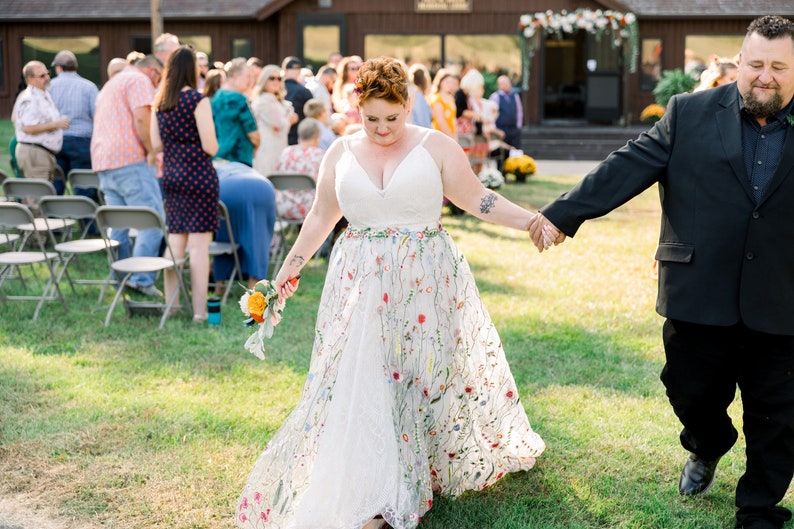 May include: A bride and groom walking down the aisle at an outdoor wedding ceremony. The bride is wearing a white dress with floral embroidery and carrying a bouquet of orange and yellow flowers. The groom is wearing a black suit and tie.